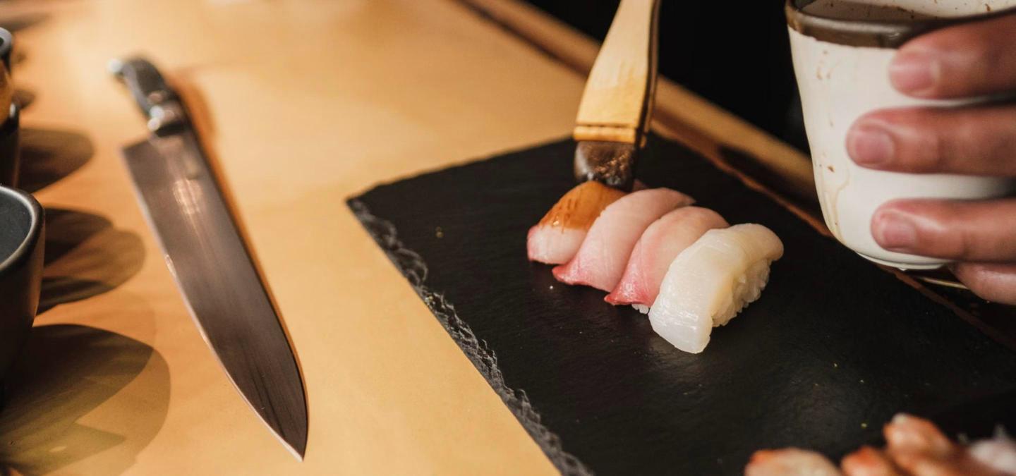 Close-up of a sushi chef's hands plating fresh nigiri at a wooden sushi counter, representing a new omakase bar.