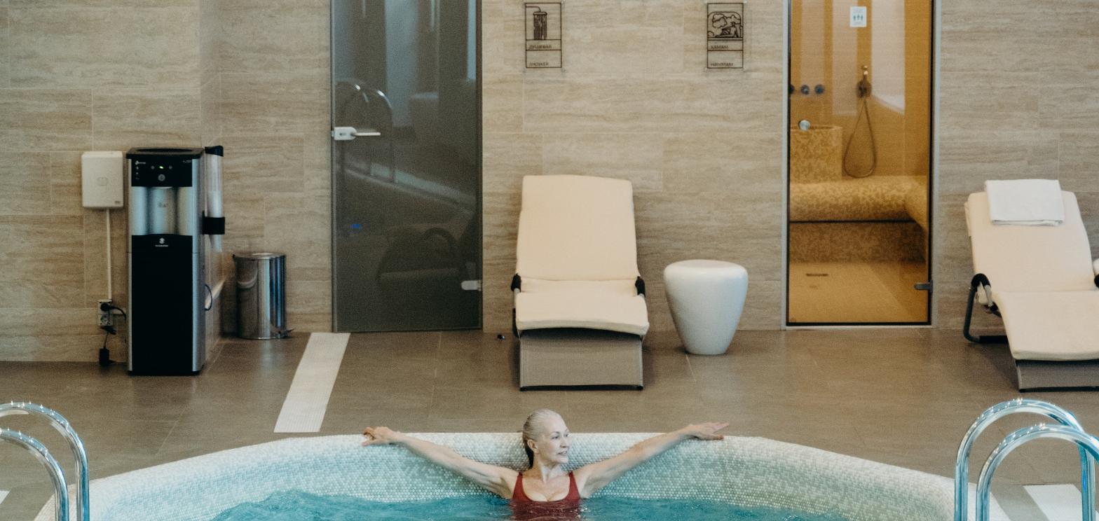 Woman relaxing in an indoor spa pool with saunas and lounge chairs in the background, representing a wellness club buildout.