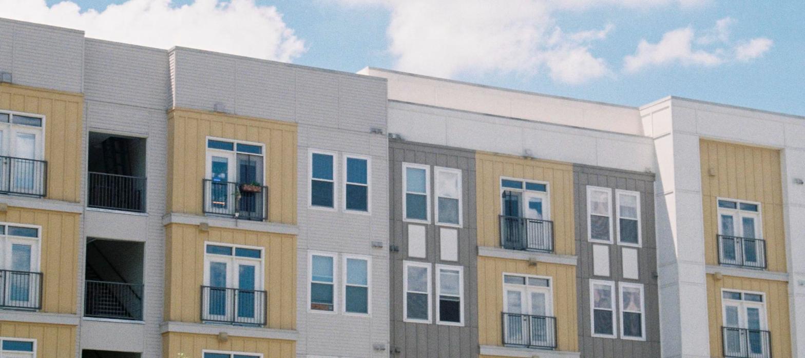 Tan and gray garden-style apartment building exterior under a blue sky, representing a suburban multifamily development.
