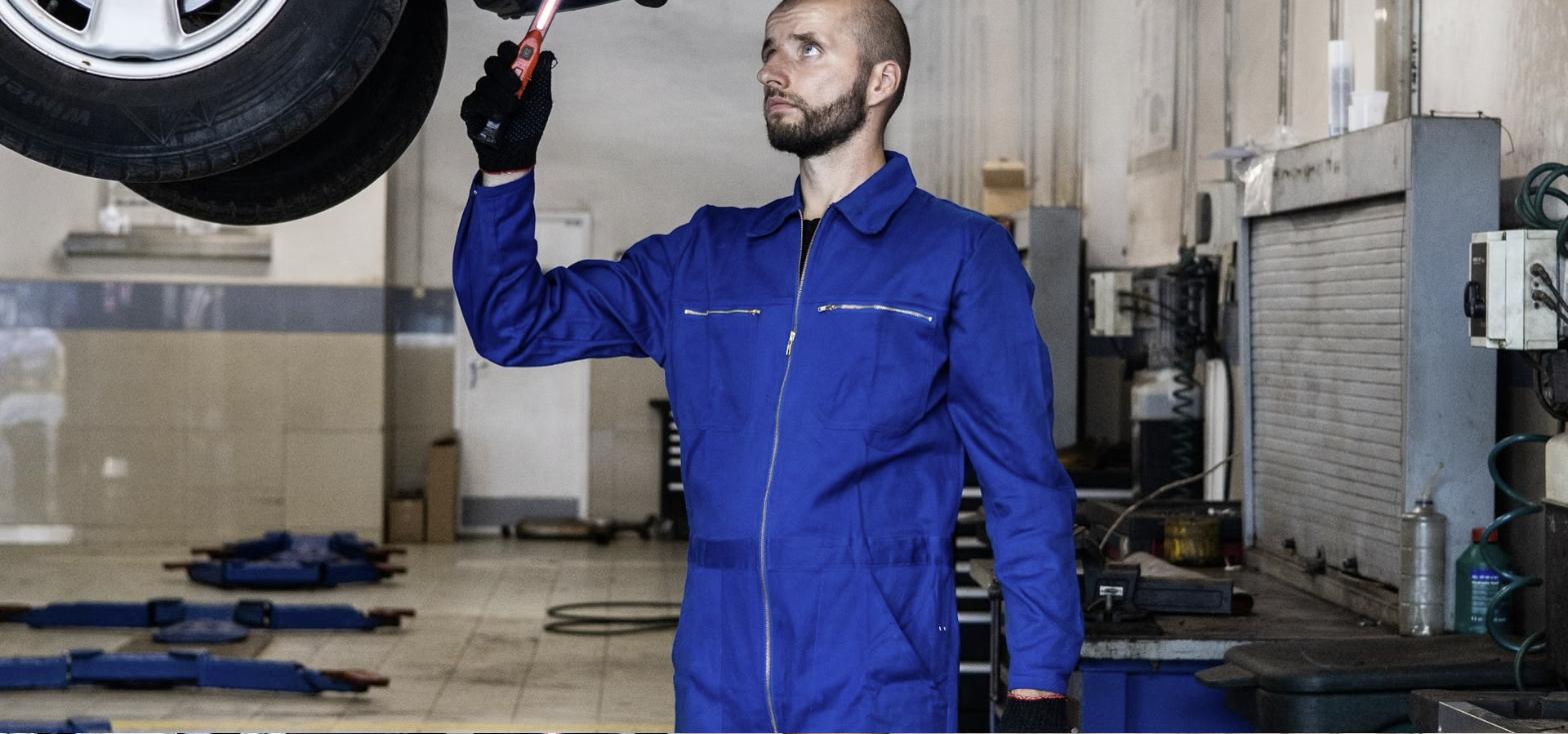 Auto mechanic in blue coveralls inspecting the underside of a vehicle on a lift in a repair garage, representing a dealership service center buildout.