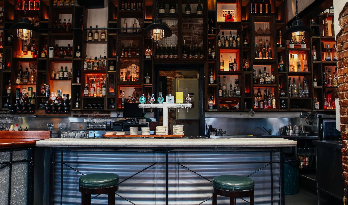 Dimly lit bar with a well-stocked back bar shelf, representing a cantina space before closing.