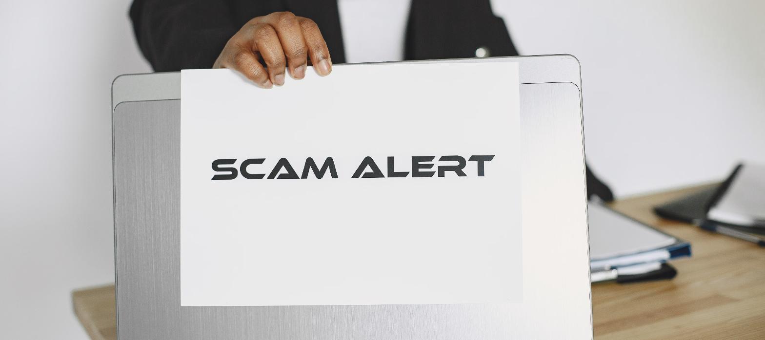 Person holding a printed scam alert sign against a laptop, representing a phishing invoice warning.